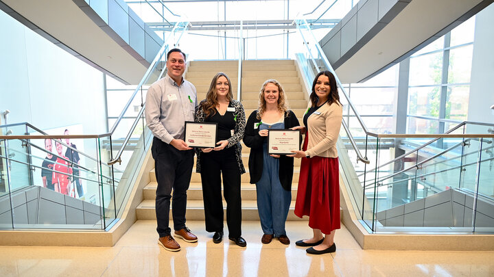 Four people standing in front of a staircase holding certificates in a bright, modern building.