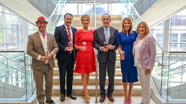 Six people standing on stairs indoors, dressed formally, each holding a glass award.