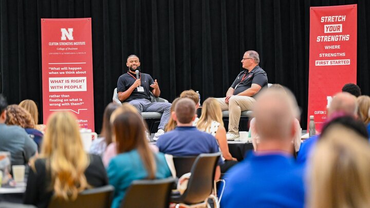 Two speakers engage in a discussion onstage with an audience seated in the foreground.