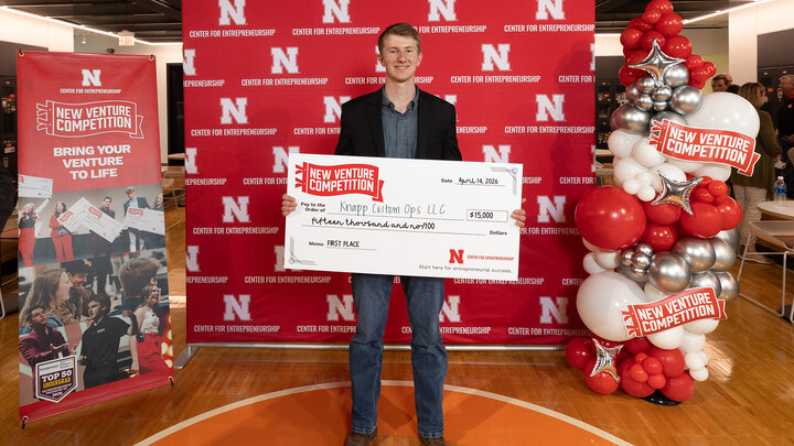 Man holding a large check at an event with red-themed banners and balloons in the background.