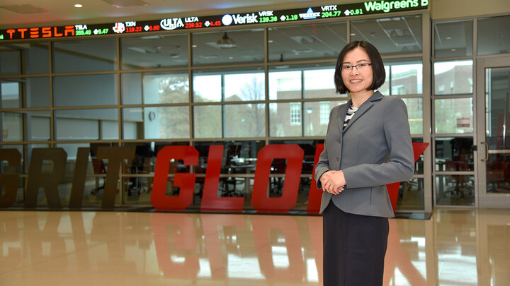 Woman stands in front of stock ticker in Hawks Hall and sign that says Grit and Glory
