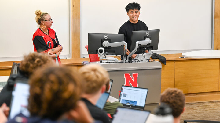 Two people speaking at a podium in a classroom, students listening with laptops open.