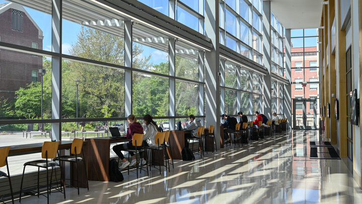 Sunny hallway with tall windows, people seated at tables, and reflections on the glossy floor.