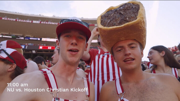 Students wear red and white striped overalls in the student section. One wears a giant Runza shaped hat.