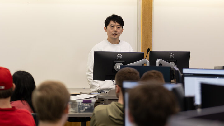 Speaker stands at the front of a classroom, facing students seated at desks with computers.