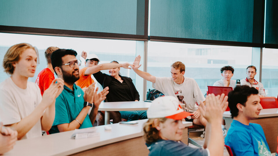 Students in a classroom, some high-fiving, others clapping, with natural light from large windows.
