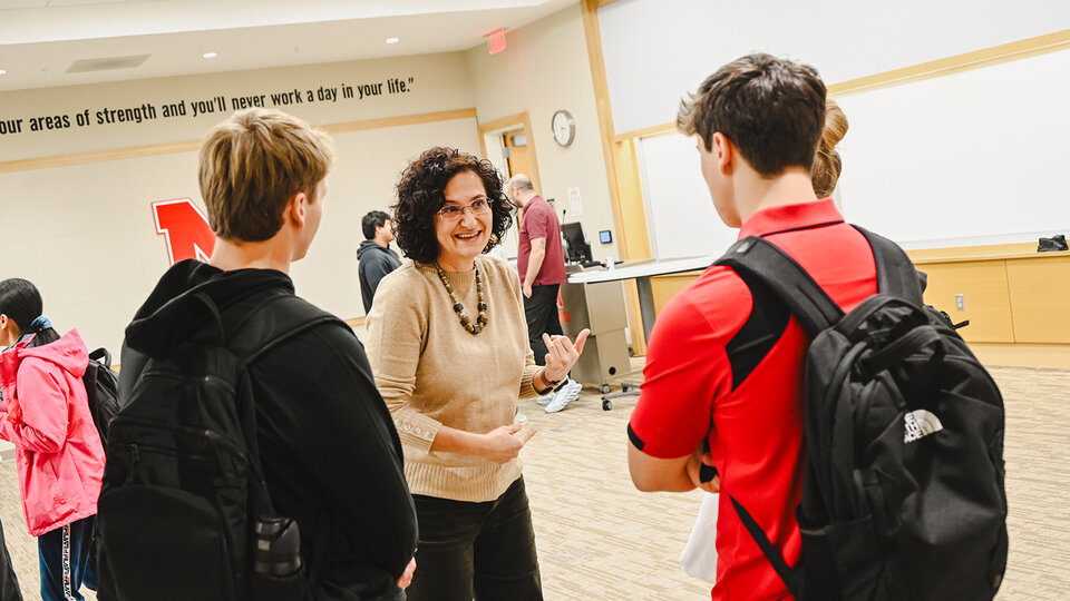 A woman speaks to two students wearing backpacks in a classroom setting.