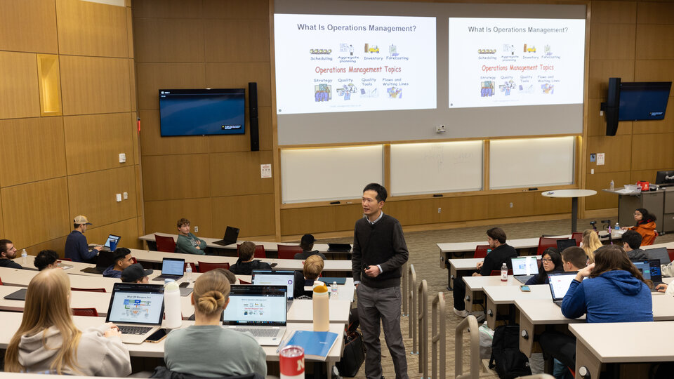 Classroom with students seated at desks and a lecturer standing near slides on operations management.