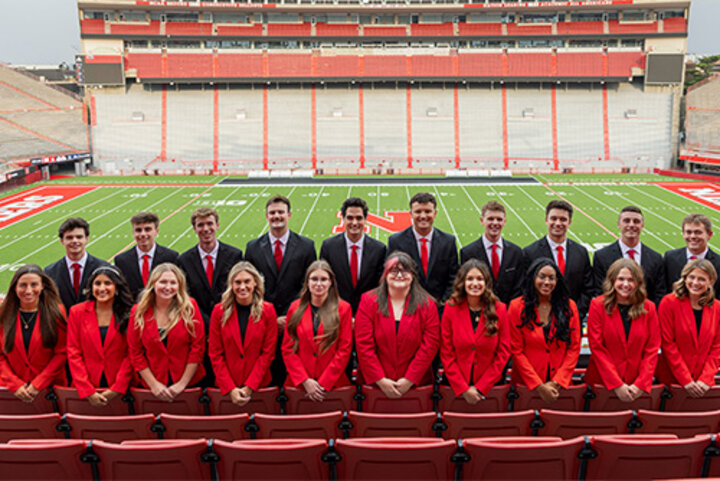 20 Huskers in red and black suits pose in Memorial Stadium