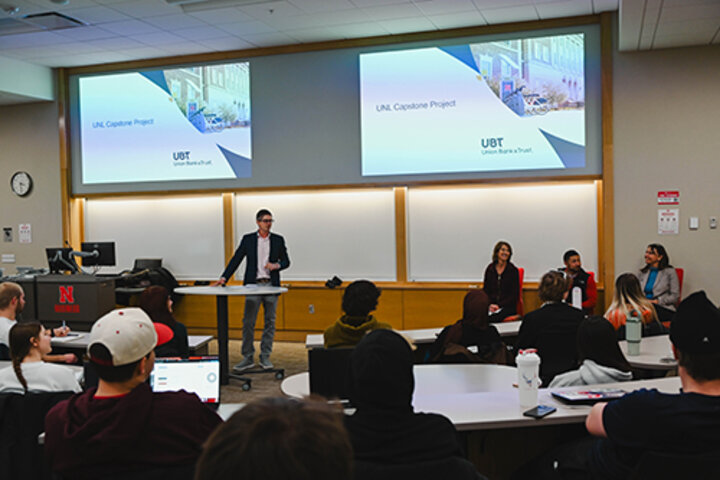 Classroom presentation with speaker, seated panelists, and audience in a university setting.