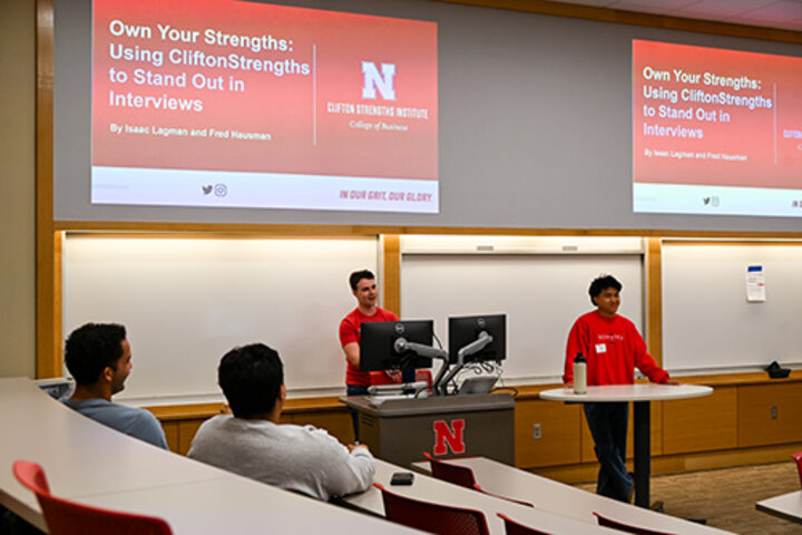 Two presenters standing at the front of a lecture room with slides displayed on two screens.