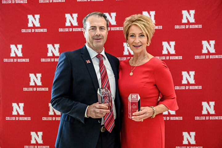Lori and Paul Hogan holding awards in front of a red College of Business backdrop.