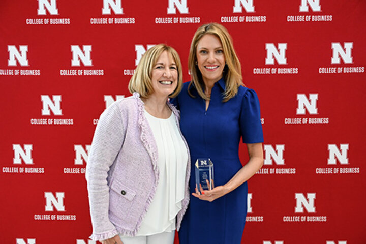 Two women standing together, one holding a glass award, on a red College of Business backdrop.