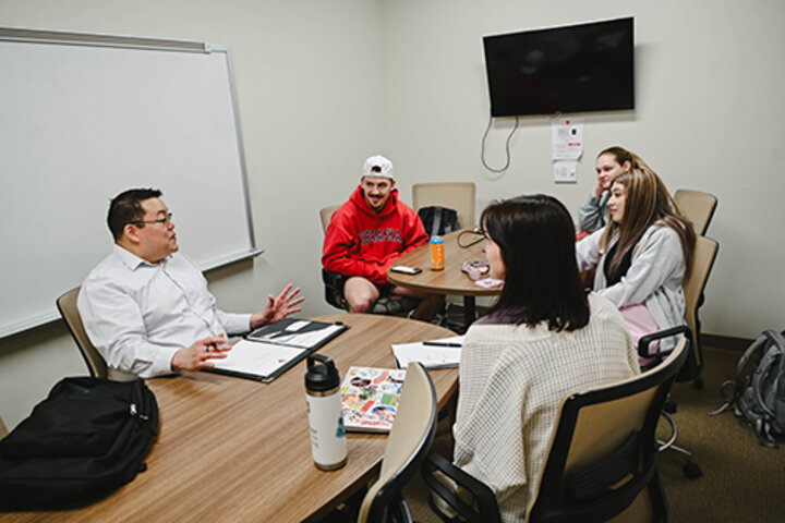 Group of four people conversing at a table in an office with a whiteboard and TV on the walls.