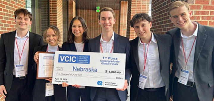 Group of people holding a large check and plaque, smiling in a brick hallway setting.
