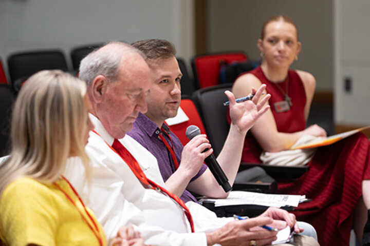 Panel discussion in a conference room, participant holding a microphone while others listen attentively.