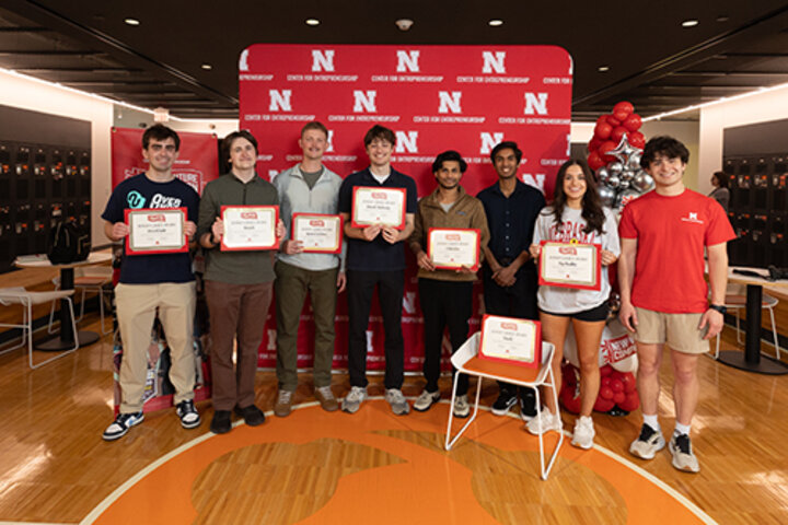 Group of people standing indoors holding certificates, with a red "N" backdrop and balloons nearby.