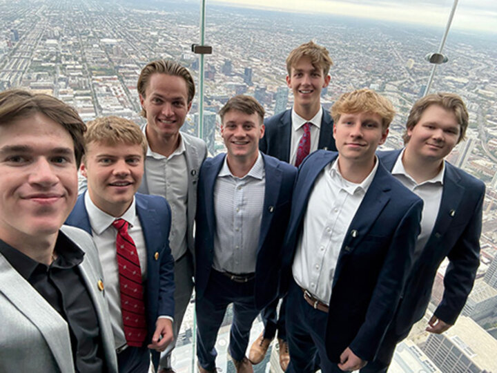 Group of people in formal attire standing on a high-rise observation deck, city landscape visible below.