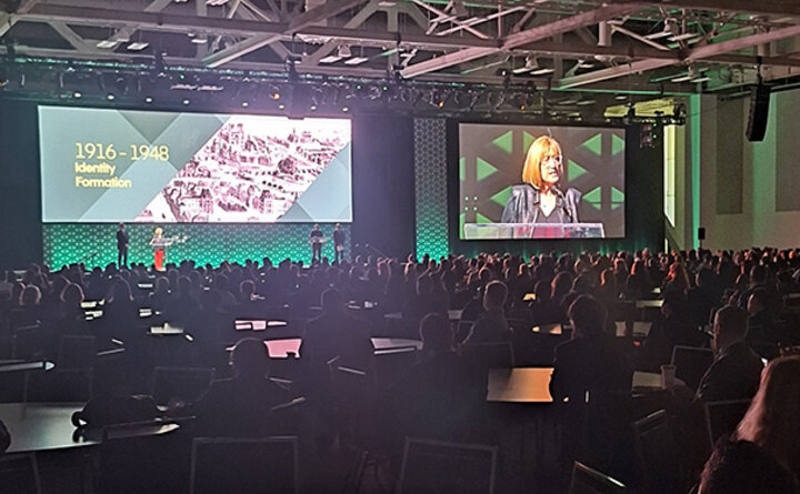 Large conference with a speaker on stage, audience seated, and slides displayed on screen overhead.