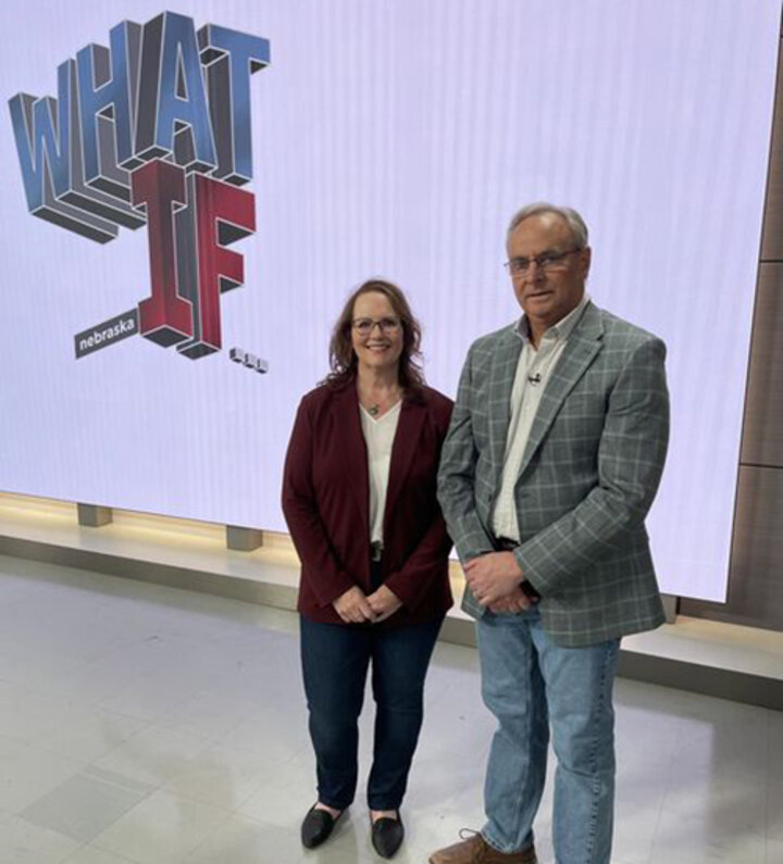 Two people standing in front of a screen displaying "WHAT IF Nebraska" in bold text.