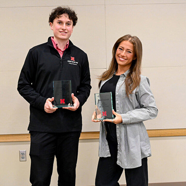 Two people holding awards, standing indoors against a beige wall, dressed in business-casual attire.