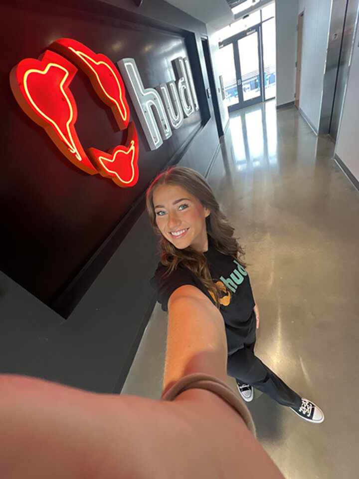 Neon hudl logo on a dark wall, person taking a selfie in a modern hallway.