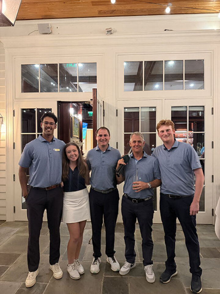 Five people in matching blue shirts pose smiling together indoors in front of glass doors.