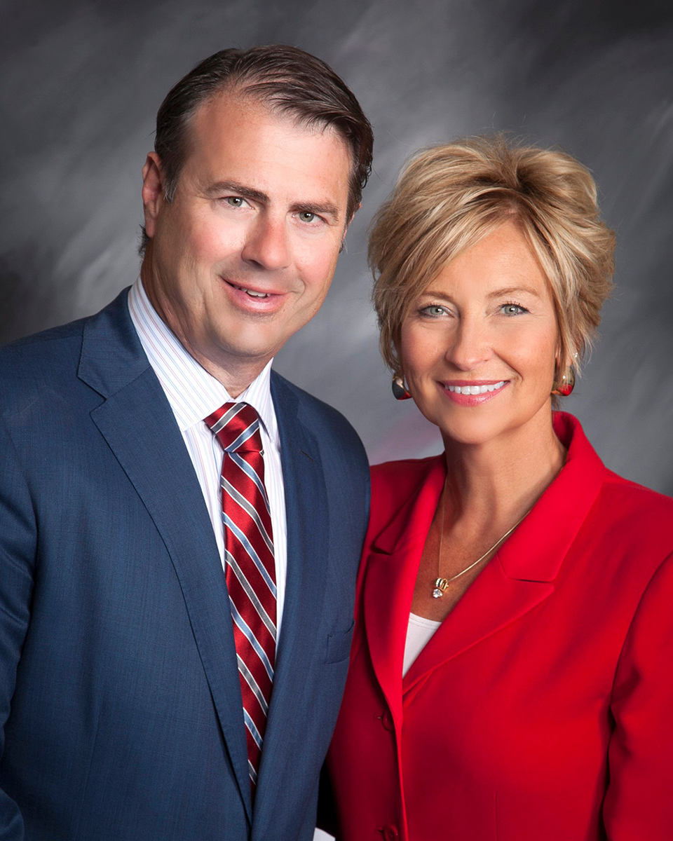 Paul Hogan in blue suit with red tie and Lori Hogan in red blazer, blurred faces, gray gradient background.